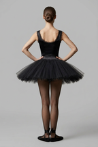 A woman from the back wearing a black ballet tutu dress with a tricot nylon bodice, black tulle skirt, sheer black tights, and black ballet flats against a light gray studio background.