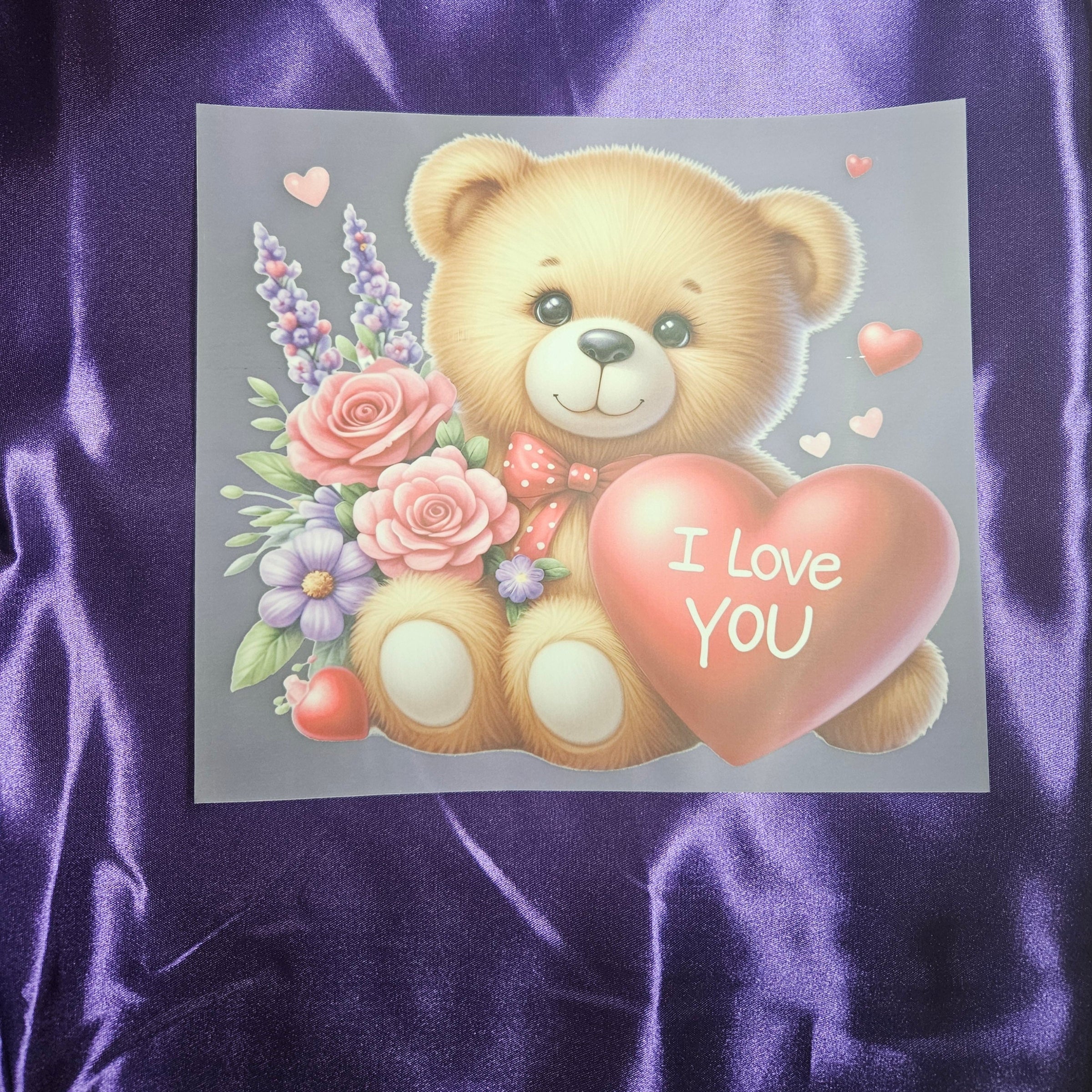 Cartoon teddy bear holding a heart with 'I Love You' text applique on a purple satin fabric background