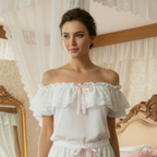 Woman in a white off-shoulder chemise standing in a bedroom with a canopy bed and decorative mirror.