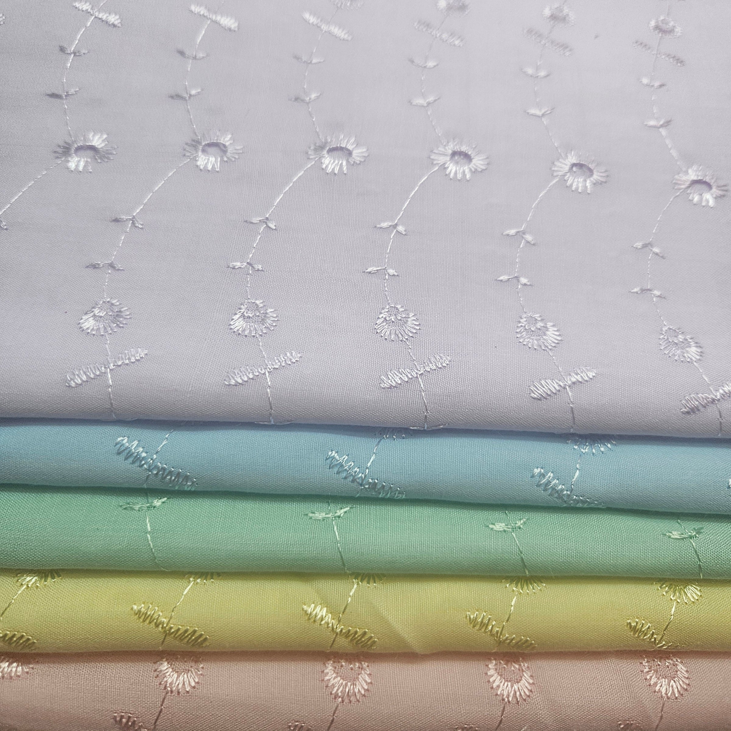 Stack of fabric swatches with floral embroidery patterns on a light lavender eyelet background.