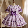 A lavender and purple floral Lolita-style dress displayed on a mannequin in a softly lit nursery setting. The dress features a large purple bow at the waist, short puff sleeves with lace trim and bows, a Peter Pan collar, and a tiered skirt with white lace trim.