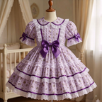 A lavender and purple floral Lolita-style dress displayed on a mannequin in a softly lit nursery setting. The dress features a large purple bow at the waist, short puff sleeves with lace trim and bows, a Peter Pan collar, and a tiered skirt with white lace trim.